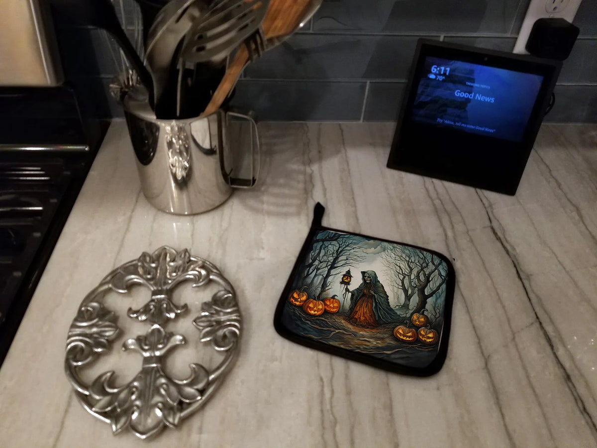 A Halloween-themed kitchen scene showing a black potholder with a printed illustration of a ghostly figure, orange pumpkins, and bare trees, alongside a silver metal trivet and a digital clock on a marble countertop.