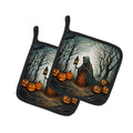 Two square potholder mats with black handles, featuring a Halloween-themed illustration of a cloaked figure in a spooky forest with glowing jack-o'-lanterns.